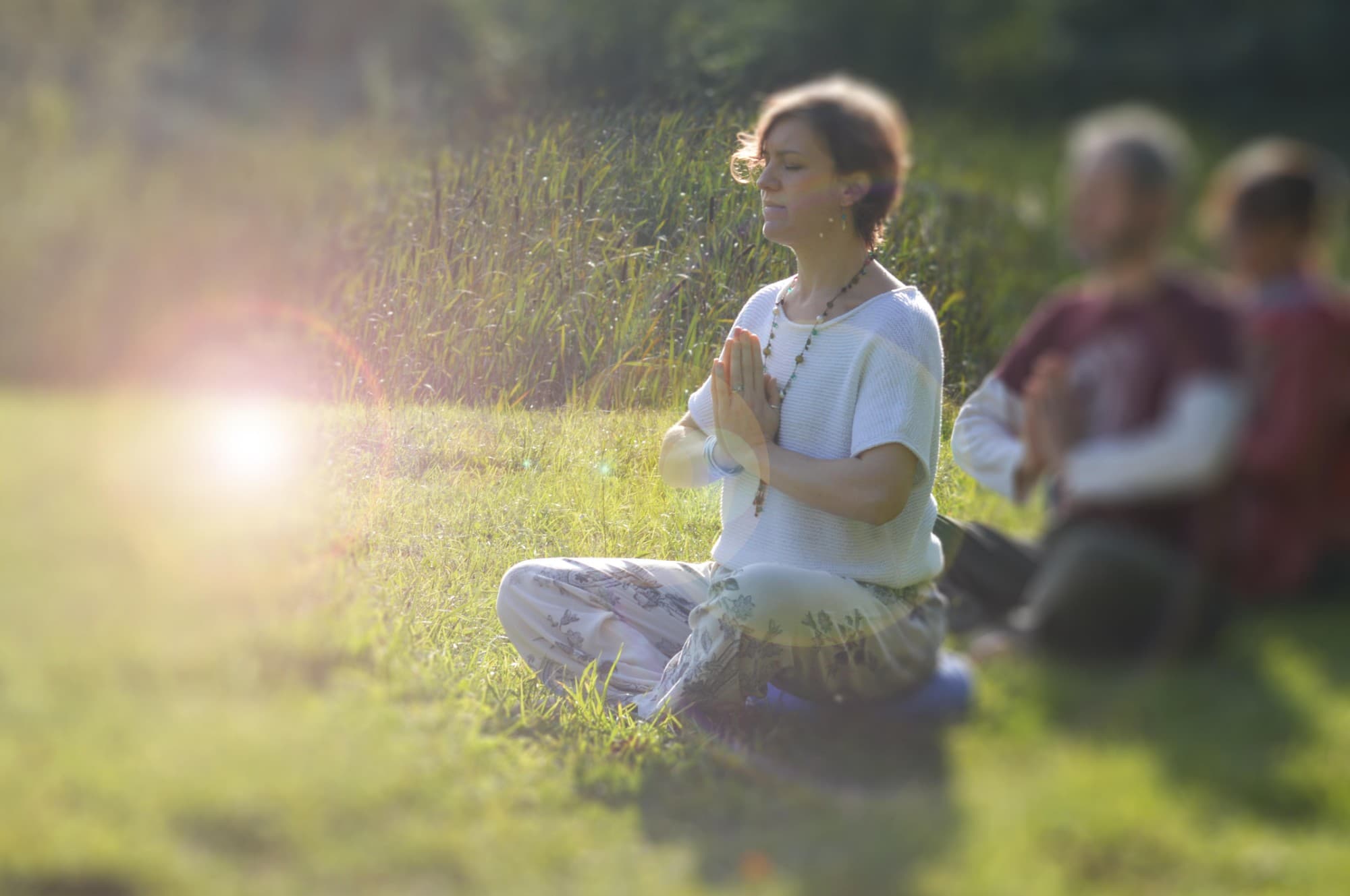 A woman meditating cross-legged in a sunlit field, hands in prayer